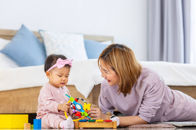 640 asian mother is playing with her pretty smiling baby daughter with wooden toy block while spending quality time in the bed for family happiness and parenting concept